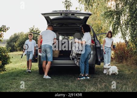 Four Members Family Traveling by Minivan Car, Mother and Father with Two Teenage Children and Pet West Highland White Terrier Dog Having Picnic Stock Photo