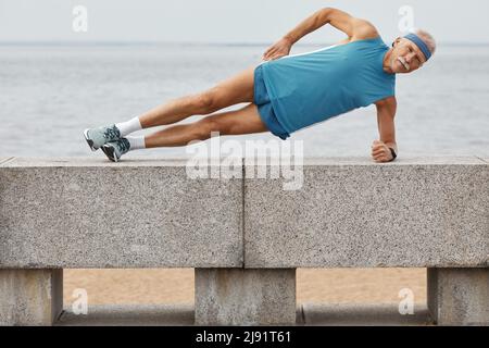 Horizontal full shot of senior man wearing sporty summer outfit doing side plank exercise Stock Photo