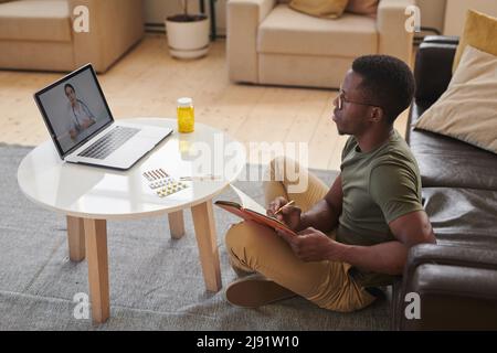 High angle shot of young adult African American man sitting in front of laptop watching doctor online and making notes while staying at home Stock Photo