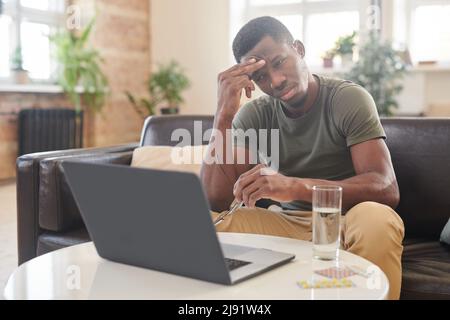 Young adult African American man sitting at desk in front of laptop holding eyeglasses suffering severe headache Stock Photo