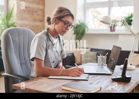 Medium portrait shot of serious female doctor wearing gray uniform sitting at desk doing paperwork Stock Photo
