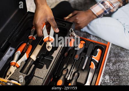 Close up of african american man in casual clothes taking pliers out of box at home. Focus on plastic black container with variety of modern tools for housework. Stock Photo