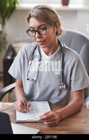 Young adult female doctor wearing eyeglasses and earphones sitting at desk in front of laptop working with patient online on video call and making not Stock Photo