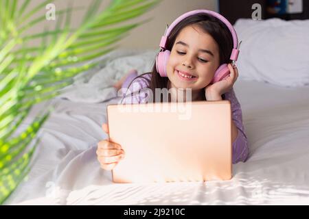 Cute little girl in pink headphones lying on a white bed, holding a pink digital tablet Stock Photo