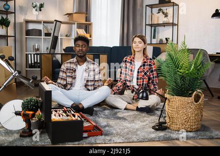 Relaxed multi ethnic couple sitting in lotus position and meditating on floor with cardboard boxes and furniture around. Young man and woman taking rest during moving process to new apartment. Stock Photo