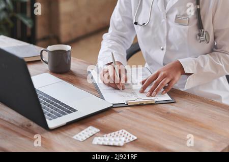 Unrecognizable female doctor wearing white coat sitting at desk in loft office writing something in papers Stock Photo