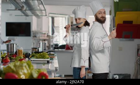 Portrait of chefs team acting funny with knives in restaurant kitchen, making joke about professional cuisine service. Cheerful man and woman working on gourmet meal dish with culinary recipe. Stock Photo