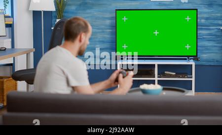 Man holding wireless controller playing console video game on green screen tv while sitting on sofa in modern living room. Gamer relaxing on couch enjoying online gaming on croma key display. Stock Photo
