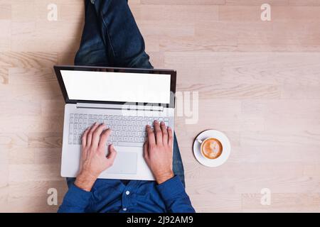 Man wearing jeans sitting on the wooden floor and holding laptop with a white empty screen Stock Photo