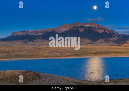 full moon setting behind sawtooth ridge and a prairie pond along the rocky mountain front near choteau, montana