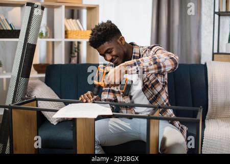 African american man sitting on cozy sofa and using cordless drill for assembling shelf at living room. Handsome young guy improving interior of his new flat. Stock Photo