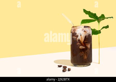 Cold coffee drink, coffee beans and leaves on a yellow isometric diagonal projection background. Can-shaped glass cup of coffee with plant-based milk. Stock Photo