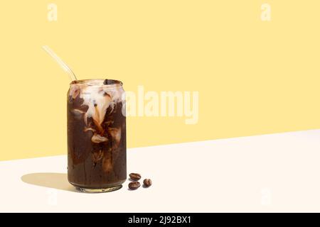 Coffee beans and cold coffee drink with glass straw on a yellow isometric diagonal projection background. Can-shaped glass of coffee with plant-based Stock Photo