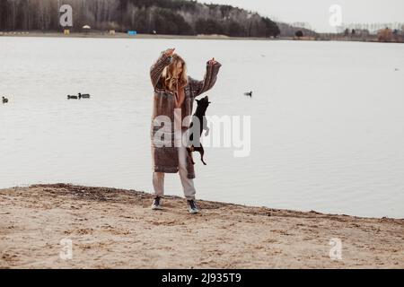 Portrait of young woman with long wavy fair hair standing on sandy beach at river, playing with small dog Zwergpinscher. Stock Photo
