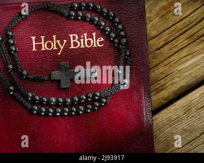 On a simple wooden table lies a Bible and a rosary crucifix. Close-up. There are no people in the photo. Solar lighting. Religious symbols, holy manus Stock Photo