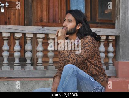 Side view of a attractive long haired Indian young man looking sideways, posing with sitting on temple stairs with his hand on chin Stock Photo