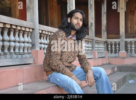 Side view of a charming long haired young man looking at camera while sitting in old temple stairs Stock Photo
