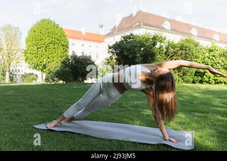 full length of slim woman practicing yoga in side plank pose on yoga mat and green lawn Stock Photo