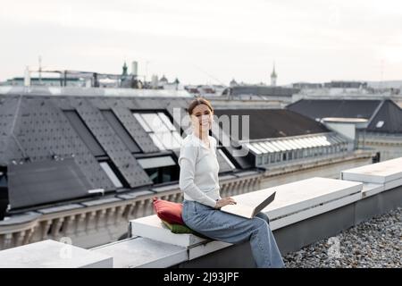 happy woman looking at camera while sitting on rooftop with laptop Stock Photo