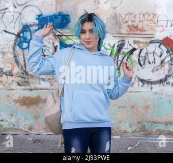 Smiling Teenager wearing light blue oversize hoodie and jeans looking into the camera. Blue haired teen girl stays outdoor against graffiti wall. Clot Stock Photo