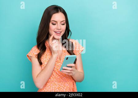 Photo of pretty tricky lady dressed orange clothes finger cheek reading app modern gadget empty space isolated teal color background Stock Photo