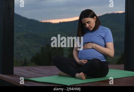 Pregnant woman hugging her tummy while sitting on yoga mat outdoor at sunset Stock Photo