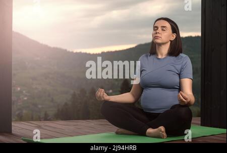 Pregnant woman practicing meditation outdoor on terrace with mountain view in background Stock Photo