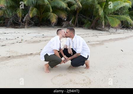 Gay couple crouching on a tropical beach drawing a heart in the sand together Stock Photo