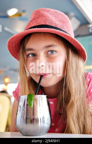 Beautiful little 10s blond girl wear summer pink hat drinks refreshing cocktail with lime through straw seated in cafe close up vertical view portrait Stock Photo