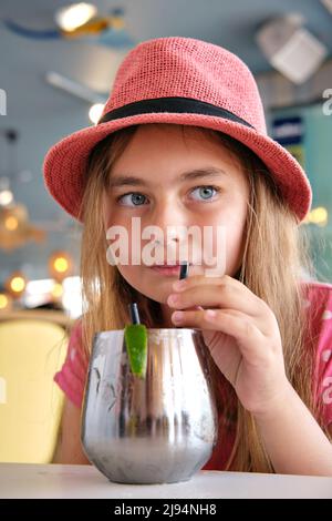 Cute little 10s blond girl wear summer pink hat drinks refreshing cocktail through straw seated in cafe close up vertical view portrait Stock Photo
