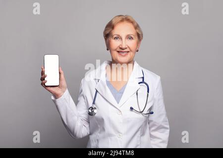 Woman doctor in white coat smiling while showing screen of his phone, using medical app, standing isolated on gray background Stock Photo