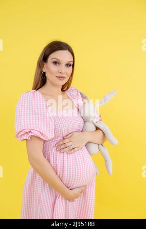 Woman with belly in late pregnancy in casual clothes smile, enjoy and hold soft toy, yellow background. Portrait of young pregnant lady expecting baby Stock Photo