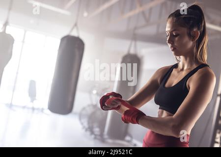 Confident caucasian young female boxer wrapping hands with red boxing wraps in health club Stock Photo
