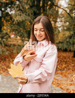 Unforgettable moments of autumn. Smiling young woman takes pictures of bright yellow maple leaf on her smartphone. She is wearing warm soft pink shirt Stock Photo