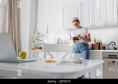 young woman writing in notebook near laptop on kitchen table on blurred foreground Stock Photo