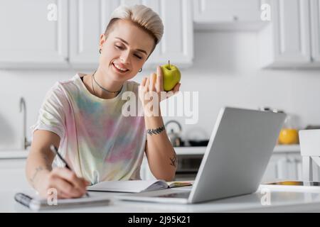 smiling woman with fresh apple writing in blurred notebook near laptop Stock Photo