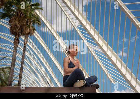 Fit woman practicing yoga and meditating outside, modern architecture and palm tree background. Stock Photo