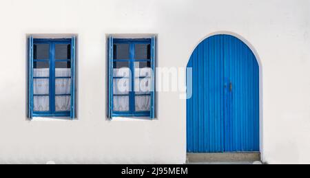 Wooden door closed and two glass windows with open shutters blue color on white wall background. Cyclades island house front view, Greek traditional a Stock Photo