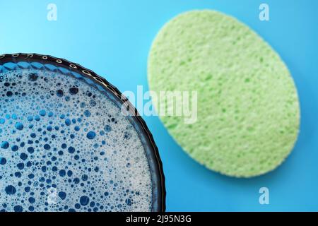 A green face sponge and a glass bowl with foamy water and bubbles bursting on a blue table Stock Photo