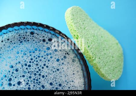 A green face sponge and a glass bowl with foamy water and bubbles bursting on a blue table Stock Photo
