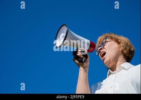 Angry senior woman in glasses shouting into a megaphone on the outside. Annoyed pensioner yells into a device amplifying sound against a blue sky Stock Photo