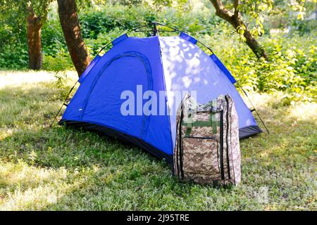 Defocus blue open tourist tent standing on green nature background. Army backpack. Tourism concept. Summer vacation in forest, camping. Lifestyle. Hik Stock Photo