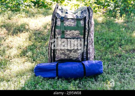 Defocus military backpack and blue tent or sleeping bag. Army bag on green grass background near tree. Military camouflage army rucksack. Vertical. To Stock Photo