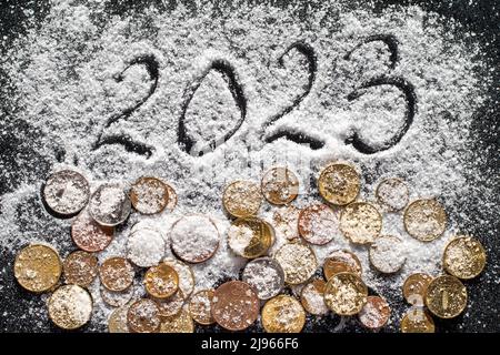 New year concept - close-up of hand written year 2023 and various coins covered with white sea salt such as snow on black background, selective focus. Stock Photo