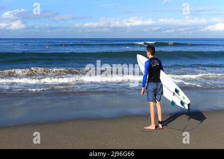 A young Caucasian surfer carrying a surfboard and standing looking out to sea at Batu Bolong beach, Canggu, Bali, Indonesia. Stock Photo