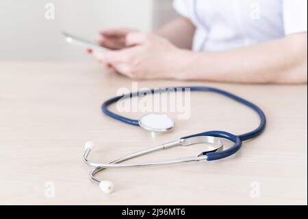 Unrecognizable female doctor sits at a desk and writes a message on a smartphone. Practitioner holds a phone in his hands and answers patients Stock Photo