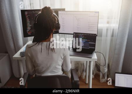 Rear of woman with box braids sitting at table coding on laptop looking at compter monitor Stock Photo