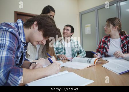 Young man writing notes during class, his classmates talking on background Stock Photo