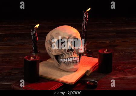 Halloween composition of spooky humans skull with burning candles. Opened satanic bible book for ritual . Stock Photo