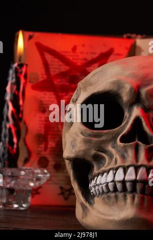 Close-up vertical shot humans skull and burning wax candle. Satanic bible on the background. Stock Photo
